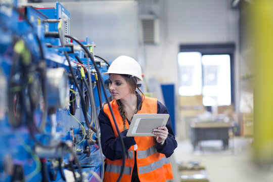 Technician with reflective vest in factory hall inspecting machine with digital tablet
