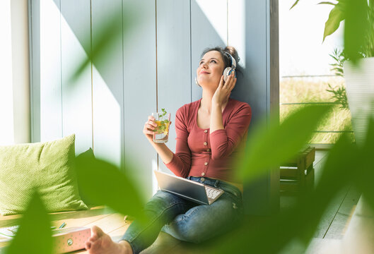 Smiling woman with headphones and laptop sitting at the window at home