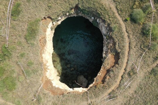Aerial view of the natural sinkhole and sea cave with turquoise water surrounded by rocky terrain and safety tape in Otranto, Apulia, Italy.