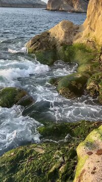 Dynamic movement of clear Atlantic water with swirling currents and foaming waves around rugged coastal rocks in warm evening light.