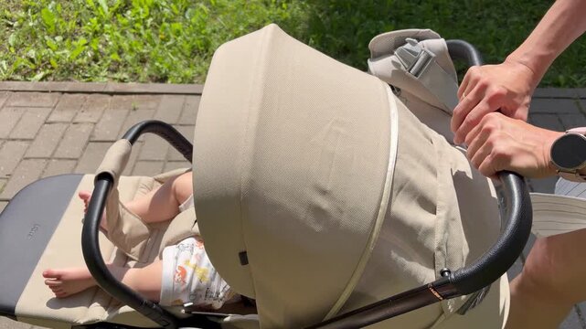 Mother pushing a beige stroller with a sleeping baby along a paved path in a park