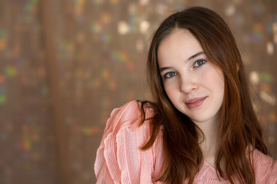 Young woman with long hair and blue eyes smiles confidently at camera in soft indoor light, close up head and shoulders portrait with warm, blurred bokeh background