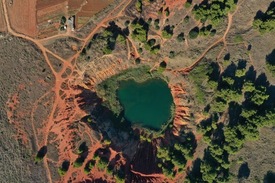 Aerial view of Laghetto Cave di Bauxite featuring its vibrant emerald green water surrounded by striking red earth cliffs and lush green vegetation in Otranto, Puglia, Italy.
