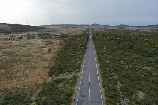 Aerial view of a person in a red jacket standing on a long straight road crossing the Paul da Serra plateau with shrubs and wind turbines in Serra de Agua, Madeira, Portugal.