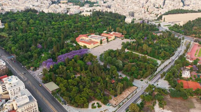 Aerial footage slowly orbiting the zappeion hall and the national garden in athens, revealing the panathenaic stadium and the dense urban cityscape surrounding the large green park