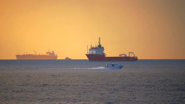 white speedboat passing in front of distant service ships outside Male harbor with orange sky dark horizon and blue gray evening water at golden hour