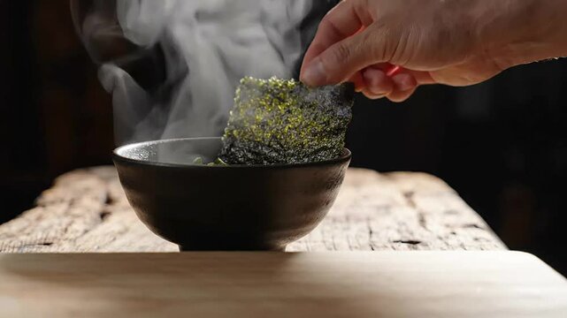 Chef hand putting nori seaweed sheet into a hot bowl of steaming miso soup on a rustic wooden table in a traditional Japanese kitchen for authentic culinary preparation