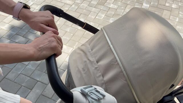 A mother's hands pushing a beige baby stroller along a paved walkway on a sunny day. A peaceful outdoor stroll in a park or city street during warm weather.