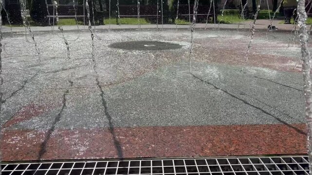 Water jets of a ground fountain splashing on red and grey granite pavement in a sunny park