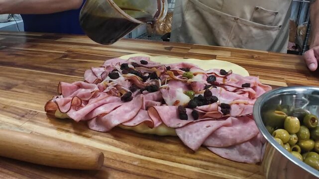 Close-up view of a professional baker drizzling sweet panela syrup over a savory ham bread dough filled with raisins and olives in a clean kitchen.