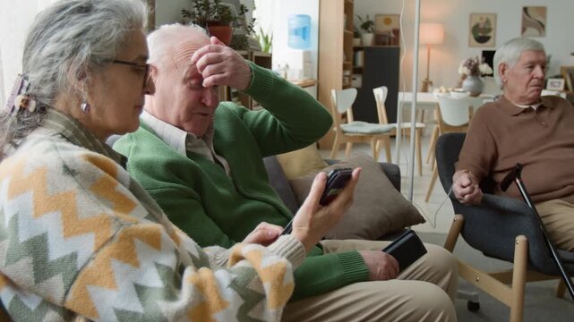 Side view shot of senior woman showing smartphone screen to senior man sitting on couch in retirement home. Clip for marketing senior-friendly apps and digital literacy