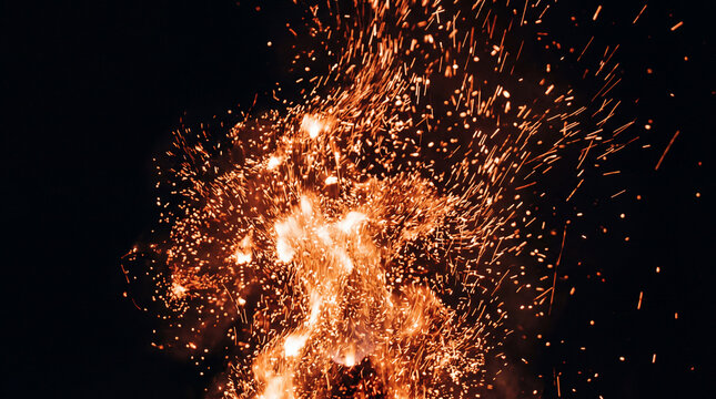 A dramatic close up of bright glowing orange fire embers and sparks flying upwards against a dark black night sky background