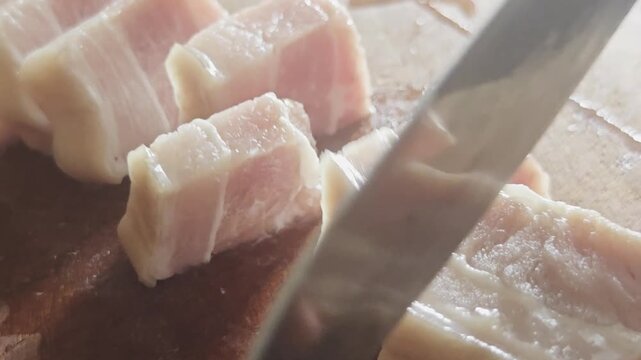 A high-quality macro close-up video capturing a person precisely slicing a fresh slab of raw pork belly into thick, uniform portions using a sharp chef's knife on a wooden cutting board