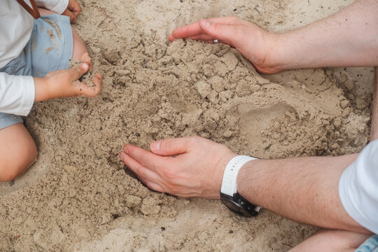 Close-up of toddler sitting on sandy beach, playing with sand beside dad
