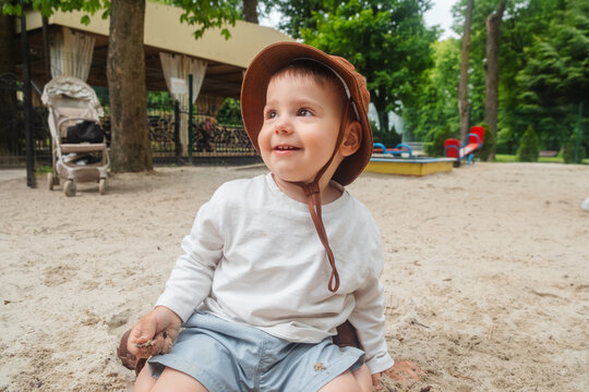 Toddler boy in brown sun hat sitting on sandy playground