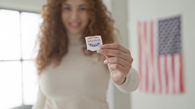 Woman holds a 'voted' sticker in hand inside a building with an american flag and blurred window; pride civic duty.