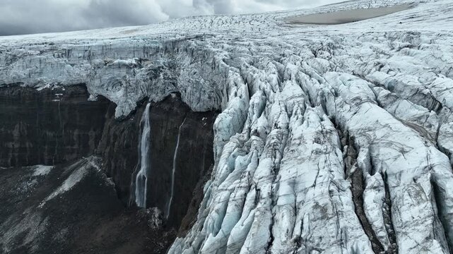 Aerial view of the glacier with deep crevasses and a waterfall cascading down the dark cliffside in Vik, Myrdalshreppur, Iceland.