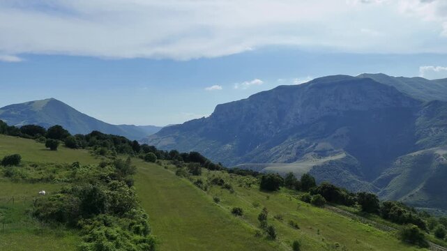 Ampio prato verde ai piedi di una montagna e cielo azzurro punteggiato da nuvole bianche. Scena naturale luminosa e tranquilla