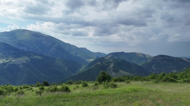 Paesaggio montano con grandi cime verdi sullo sfondo, prato erboso in primo piano e cielo parzialmente nuvoloso. Scena naturale ampia, luminosa e incontaminata
