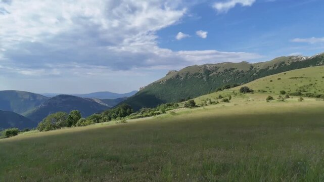 Ampio prato verde ai piedi di una montagna e cielo azzurro punteggiato da nuvole bianche. Scena naturale luminosa e tranquilla