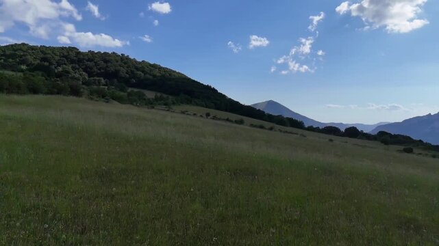 Ampio prato verde ai piedi di una montagna e cielo azzurro punteggiato da nuvole bianche. Scena naturale luminosa e tranquilla