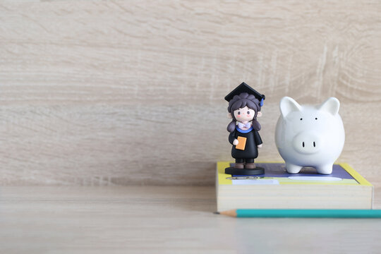Cute graduation student figurine with a piggy bank on a book, representing education savings, financial planning for children's future and academic goals.