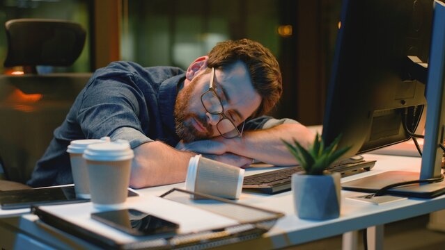 Tired Caucasian business man sleeping near computer with empty cups of coffee at night office exhausted businessman relax male employer sleep napping at dark evening company deadline problem burnout