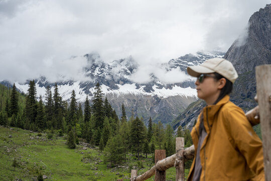 The female traveler stands with her back against a fence, with snow-capped mountains and a forest in the background