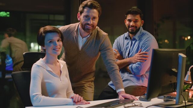 Happy business team posing at table with computer smiling looking at camera at dark office. Group of three businesspeople work talking joyous smile cooperation teamwork partnership at evening agency