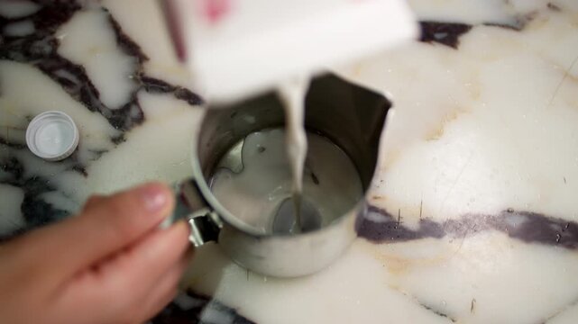 Hand pours milk from carton into metal pitcher on marble countertop, kitchen prep