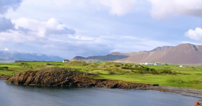 A scenic view of a vast green golf course on a rocky coast with a bay, large mountains, and a cloudy sky in the background, likely in Iceland.