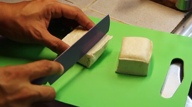 Hands slicing fresh tofu on a chopping board.