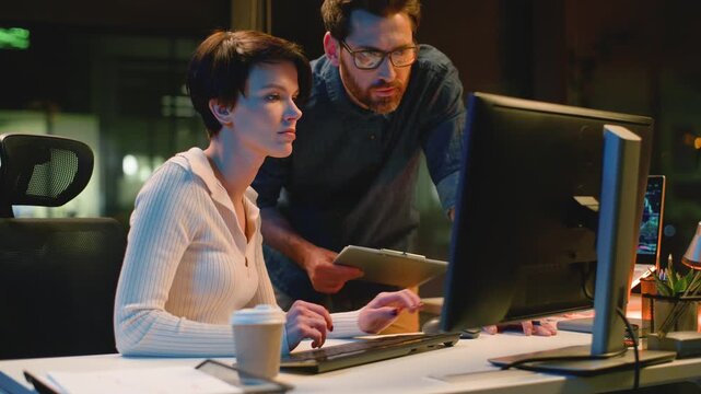 Business team partners with computer discuss work at night evening office. Caucasian man leader coming checking working process help with difficult task woman intern asking talk with boss cooperation