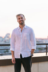 Fototapeta premium Smiling handsome young man in white linen shirt standing on rooftop terrace at sunset