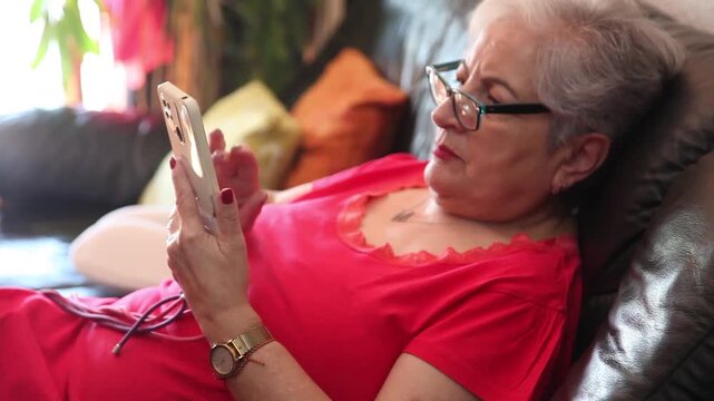  Mujer mayor con bata rosa usando un tel&eacute;fono inteligente en el sof&aacute; de su casa. Mujer mayor con gafas navegando por el tel&eacute;fono inteligente en casa. Senior activo, estilo de vida tecnol&oacute;gico moderno.