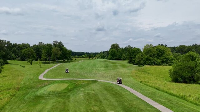 Cinematic drone shot capturing two electric golf carts changing directions on splitting paths. The footage showcases a vast green fairway surrounded by dense trees under a cloudy sky.