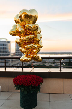 Large bouquet of red roses in box and gold heart shaped balloons on rooftop terrace at sunset