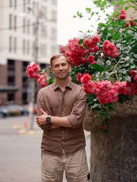 Smiling handsome man standing with arms crossed near blooming pink rose bush on city street