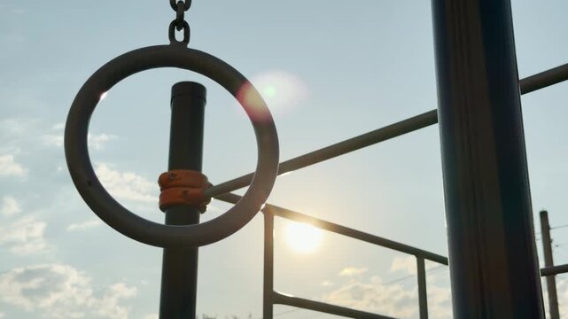A gymnastic ring hangs peacefully at an outdoor fitness park. The bright sun glows warmly behind the sturdy workout bars. This hopeful scene feels completely calm and motivating.
