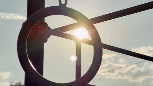 A metal playground ring hangs from a structure under a bright sky. The warm afternoon sun glows directly through it, inspiring a hopeful and serene feeling. The quiet scene feels nostalgic.