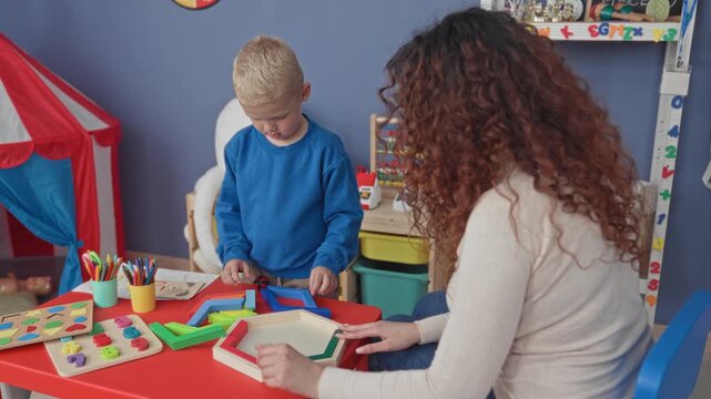 Woman guiding colorful wooden puzzle pieces on a small red table with a little boy in a playroom building; nurturing learning.