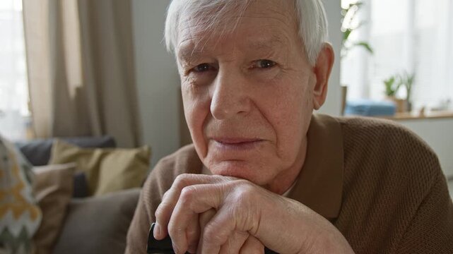 Close up portrait of senior man leaning chin on hands resting on walking cane, looking directly at camera. Footage for senior care services or retirement home advertisements