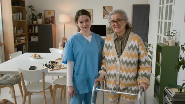 Portrait of young female nurse in blue scrubs standing next to senior woman with walker in retirement home. Footage ideal for senior care and assisted living marketing