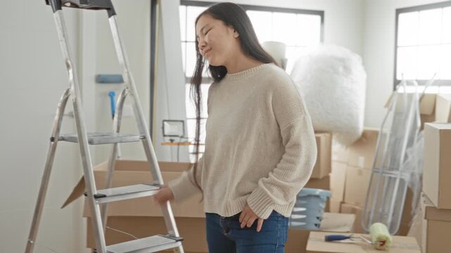 Woman holding moving boxes by a metal ladder in a building, smiling while packing and arranging taped cartons and household goods; new home joy.
