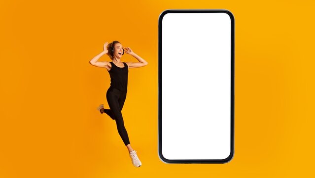 Happy Young Woman Jumping With Excitement While Holding Smartphone With Empty Screen At Yellow Studio Background. Perfect For Mockup And Advertising Concept.