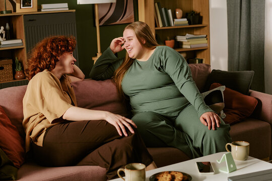 Young plus size women sitting on sofa, talking and laughing during home visit, showing friendship, emotional support, body positivity. Ideal for wellness, community, lifestyle marketing