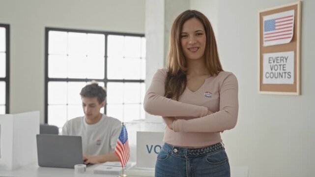 Woman with crossed arms and 'i voted' sticker stands by ballot box and small us flag while man works at laptop in polling station building; civic engagement pride.
