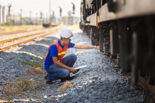 Railway inspector checking freight train suspension and wheel assembly at railyard, Engineer in hardhat and safety vests performing maintenance inspection on cargo train equipment