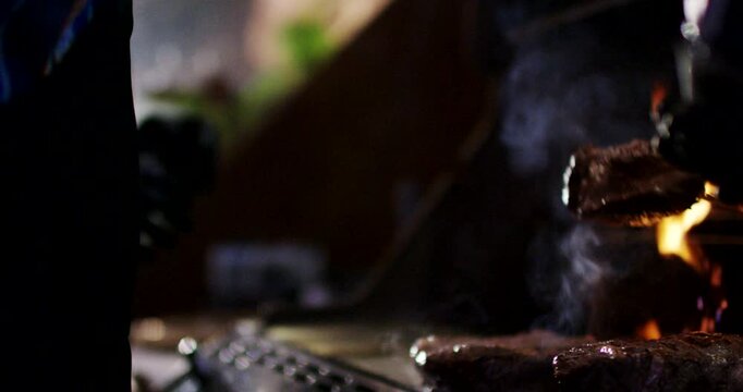 Close up view of hand using tongs to lift cooked steak from hot barbecue grill with rising flames