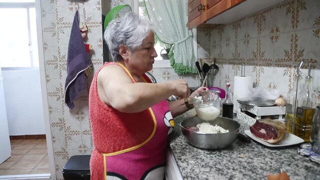  Mujer mayor agrega pan empapado en leche al taz&oacute;n para mezclar alb&oacute;ndigas. Abuela mayor agrega pan empapado en leche al taz&oacute;n. Receta tradicional de alb&oacute;ndigas caseras, aut&eacute;ntica cocina senior.